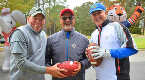 three middle-aged men dressed in college football attire at a football tailgate holding footballs