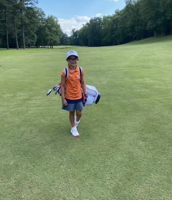 little girl in an orange shirt walking across the fairway on a golf course carrying her golf bag