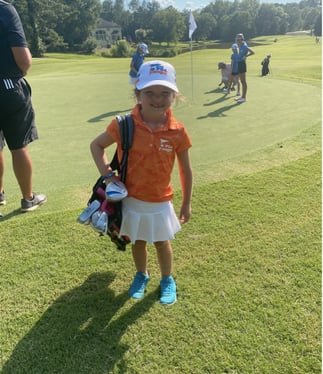 little girl in an orange shirt standing next to the green on a golf course with her golf bag